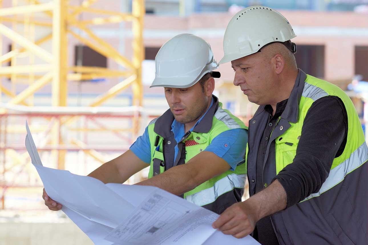 Two construction workers wearing helmets and safety vests review blueprints at a building site.