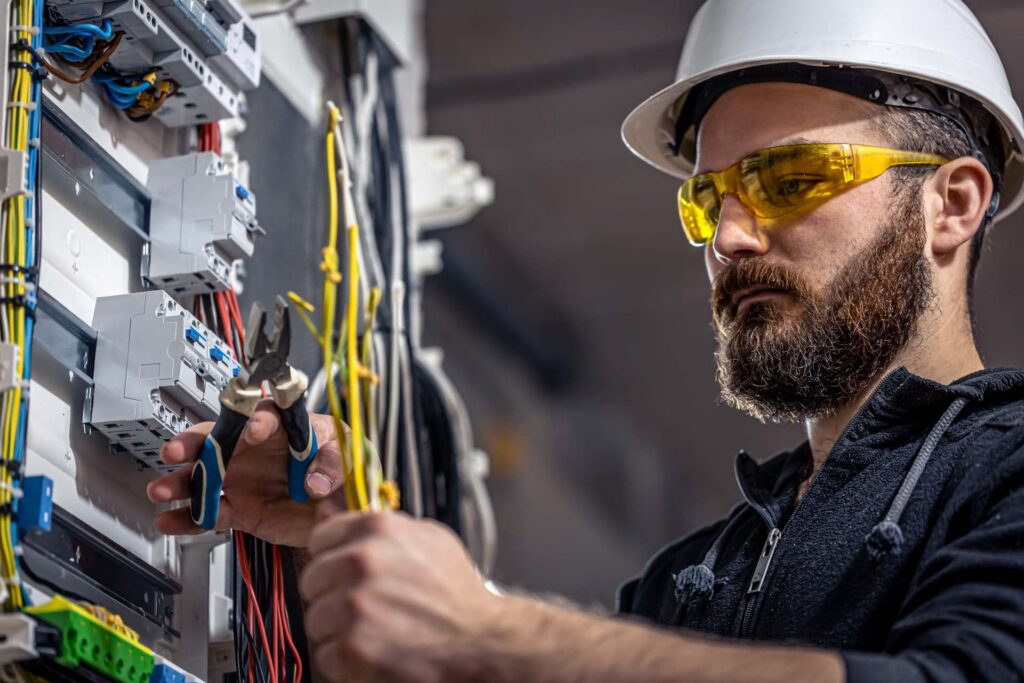 A technician wearing safety glasses and a helmet working on electrical wiring and components.